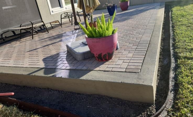 A newly constructed brick patio with a concrete border, a pink potted plant, and a grassy yard. A newly constructed brick patio with a concrete border, a pink potted plant, and a grassy yard.