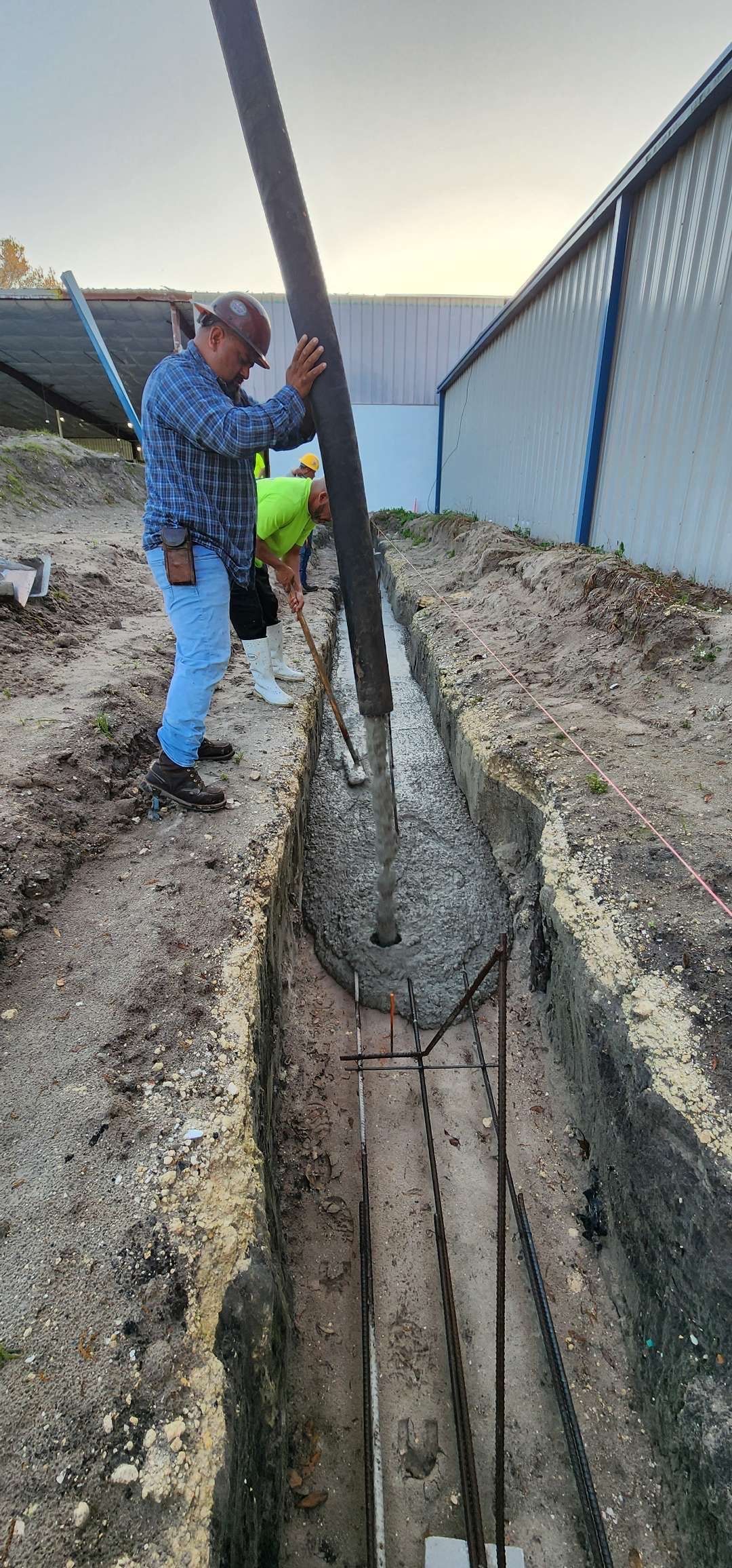 Two construction workers pouring concrete into a trench. They are next to a building.
