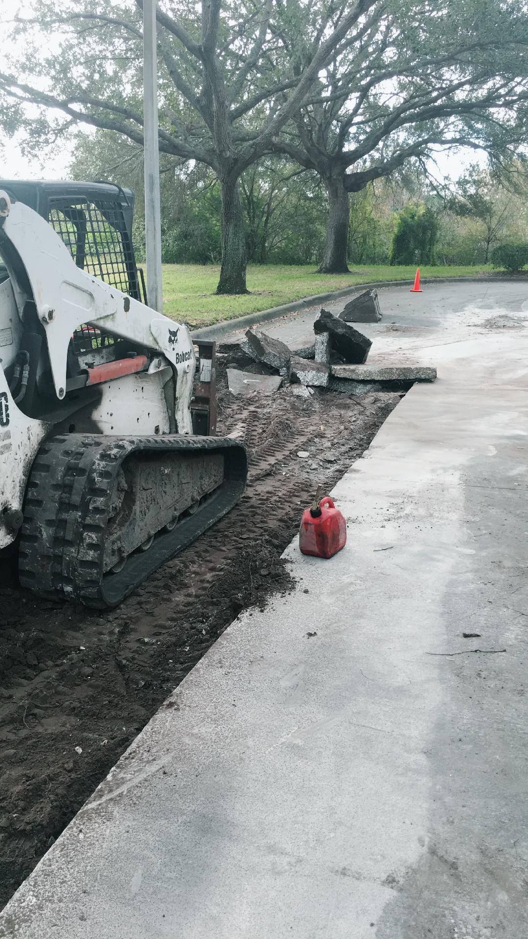 A white skid steer removing asphalt from a concrete curb. A red gas can sits nearby.