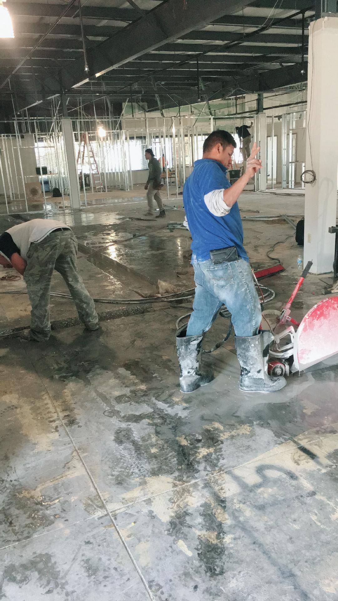 Workers in a construction zone: one using a power tool, one leaning over, and another walking. Concrete floor, indoor setting. Workers in a construction zone: one using a power tool, one leaning over, and another walking. Concrete floor, indoor setting.