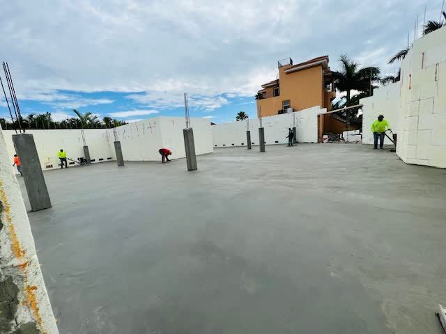 Construction site with workers pouring concrete floor. Gray concrete, white walls, cloudy sky.