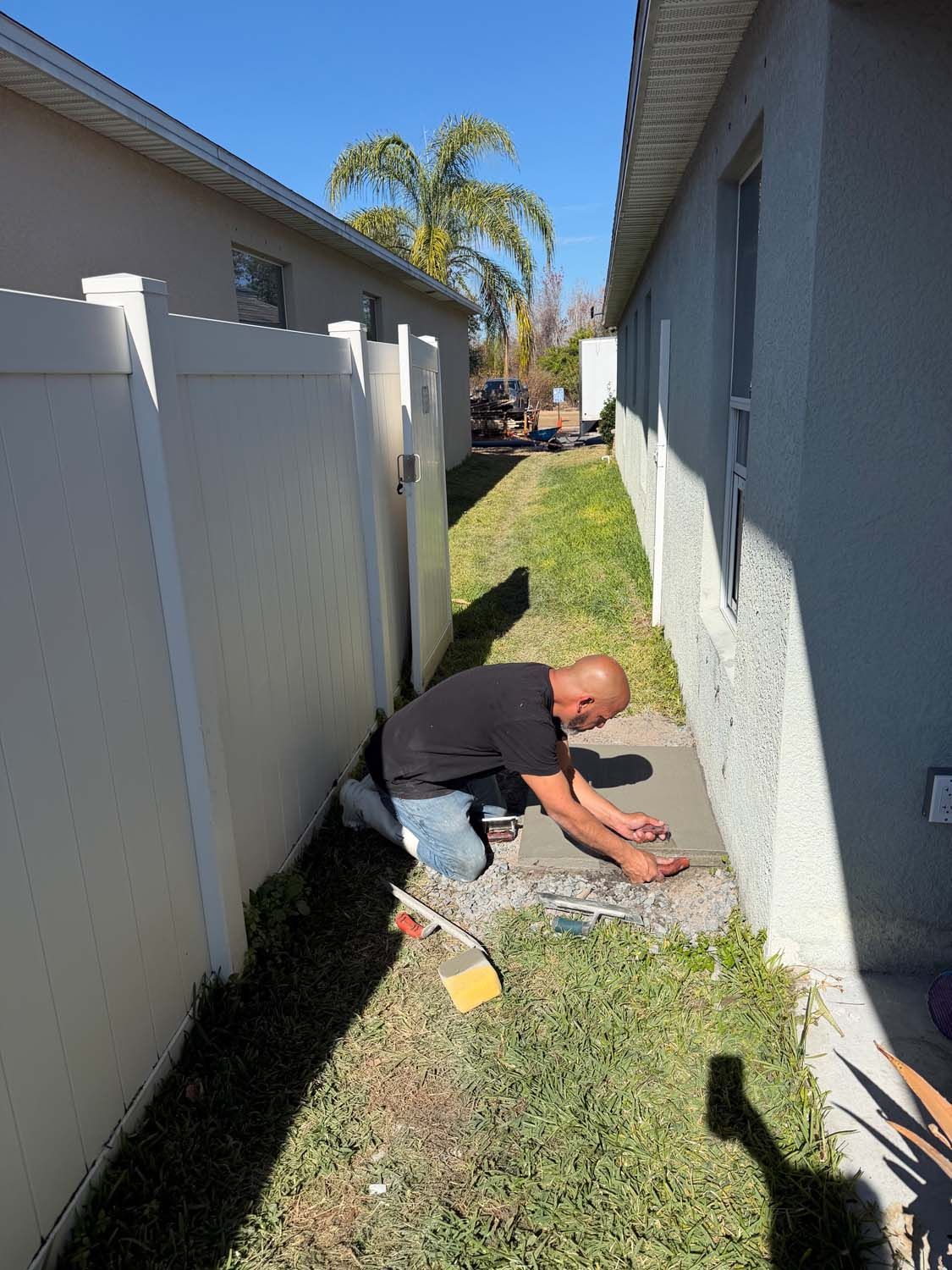 Man kneeling on a narrow strip of concrete between two buildings, working on a surface, sunny day.