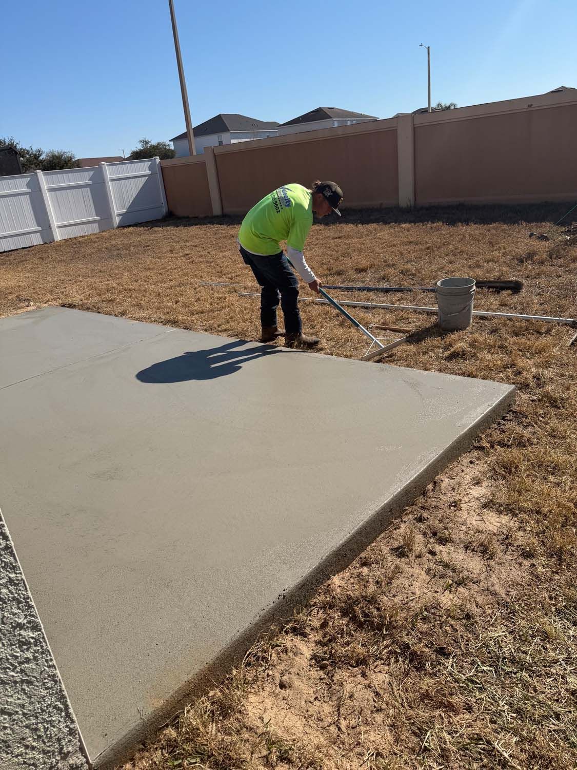 A person in a lime green shirt smoothing wet concrete with a tool in an outdoor setting.