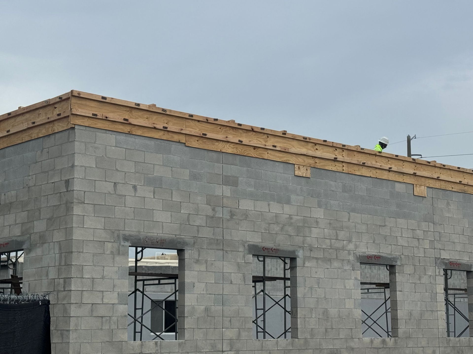Building under construction with concrete block walls, wooden roof framing, and scaffolding. A worker is visible on the roof.