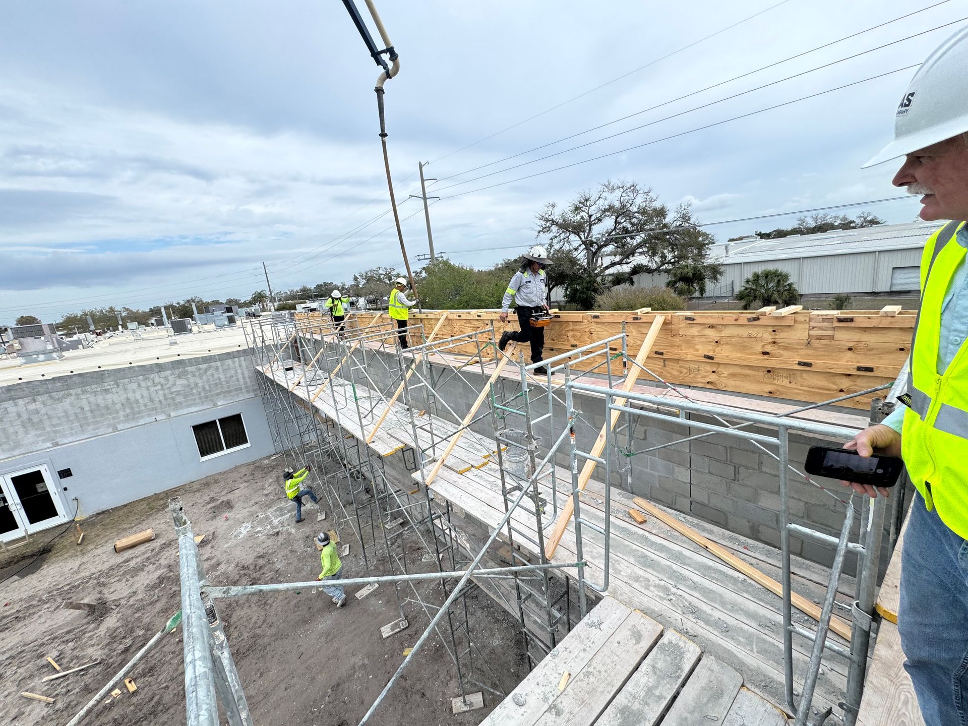 Construction site: Workers pour concrete on roof with pump. Scaffolding, safety vests, overcast sky.