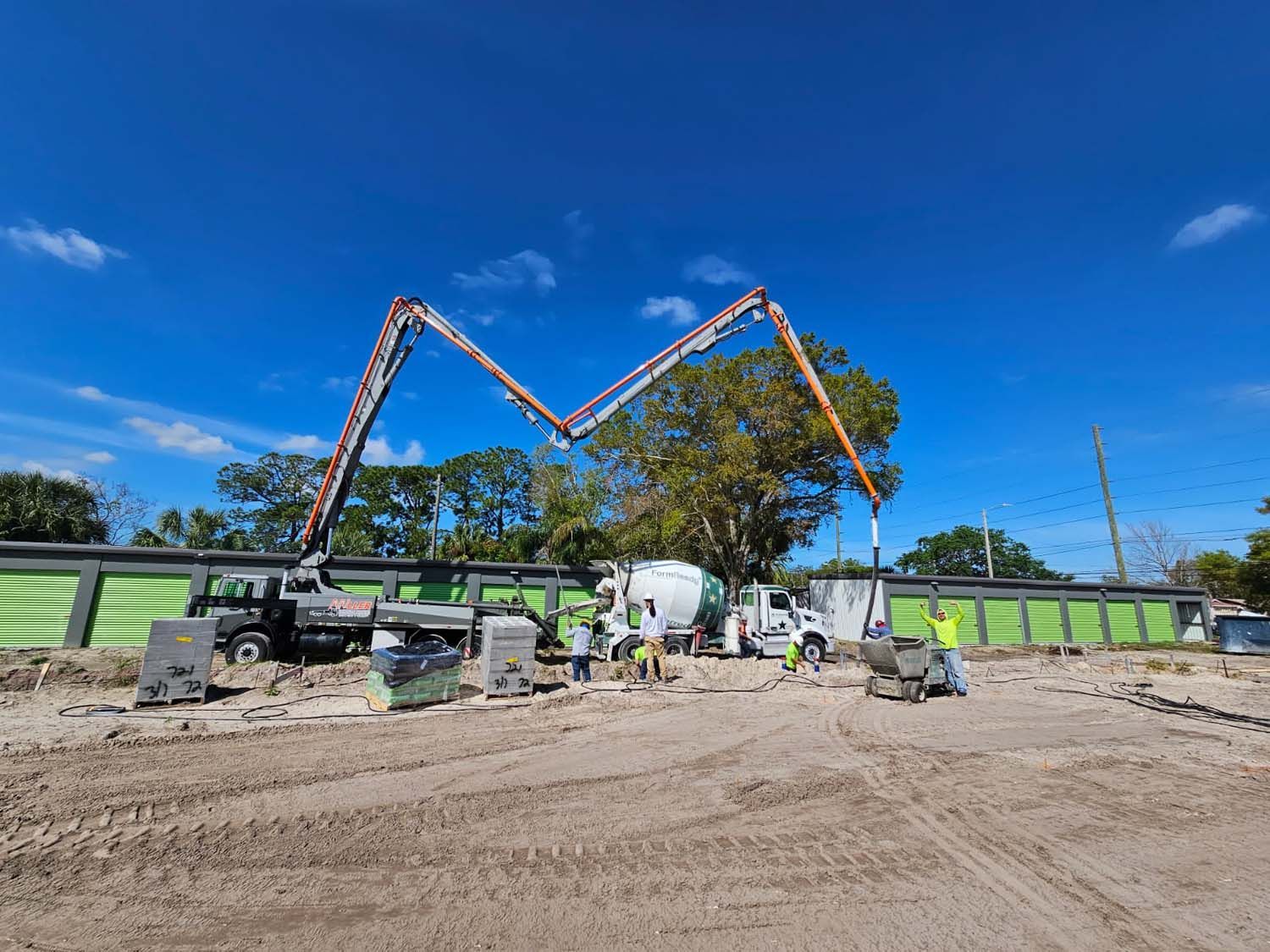Concrete truck with boom pouring concrete, storage units in background, sunny day.
