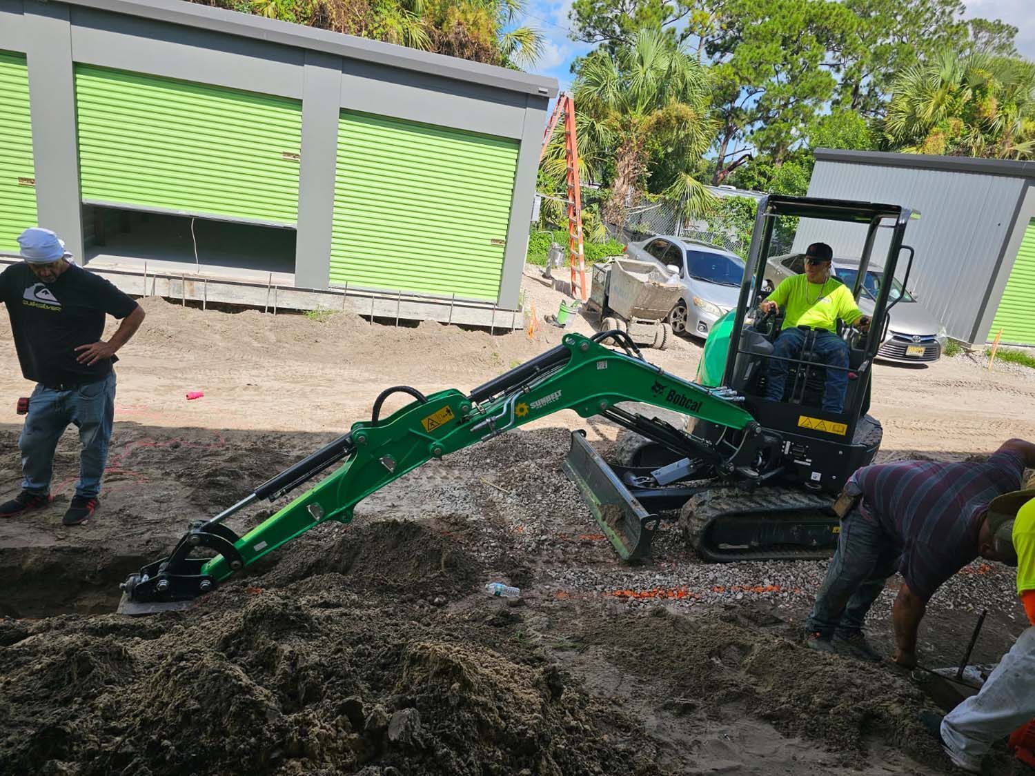 Green excavator digging in dirt; workers nearby. Green storage buildings in background.