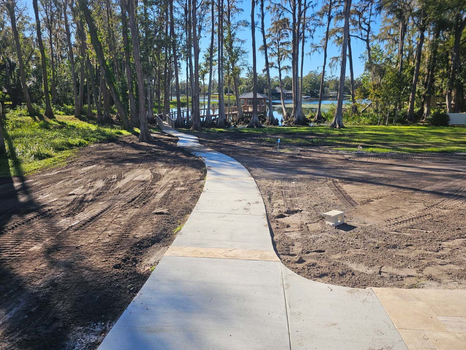 Concrete path leading towards a lake, surrounded by dirt and trees on a sunny day.
