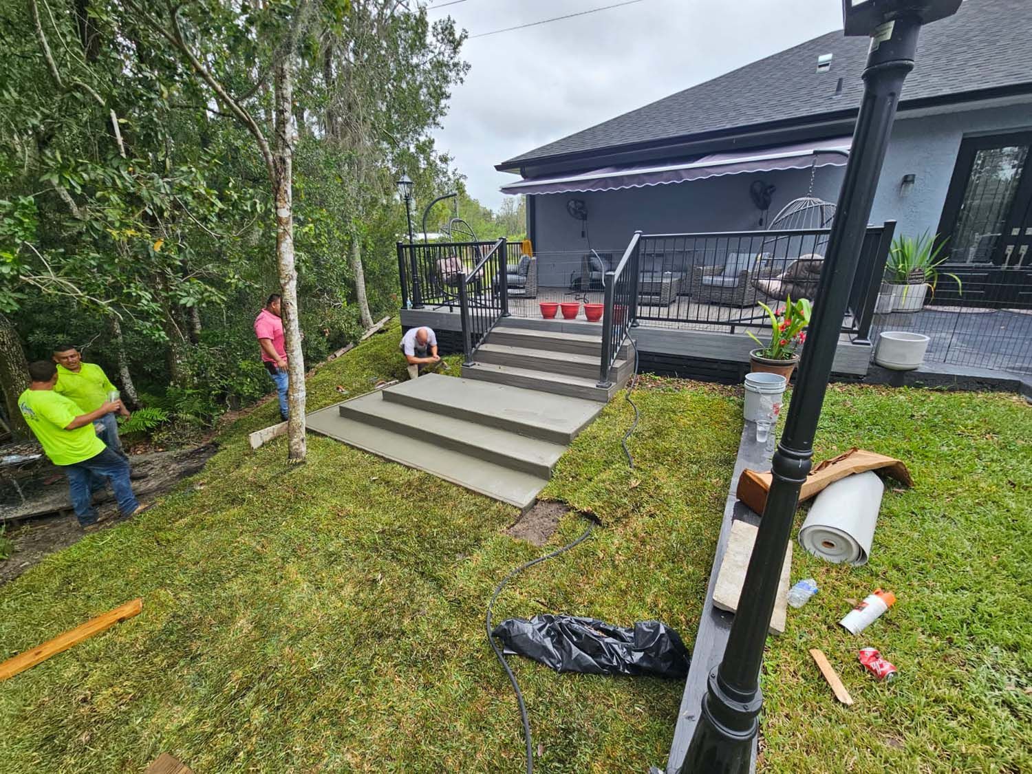 Workers installing concrete steps leading to a raised deck with black railings; lush green grass surrounds.
