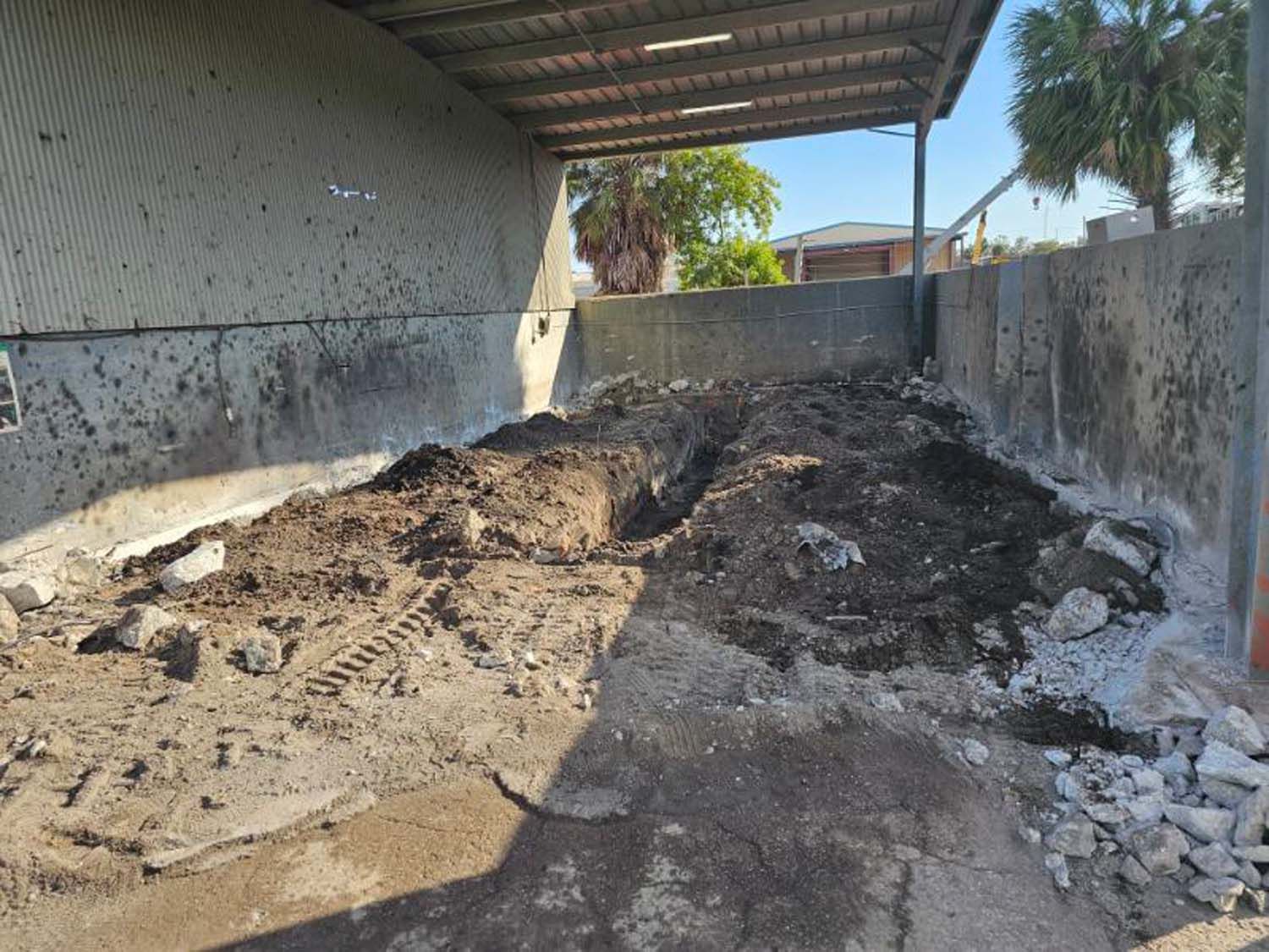 A dirt and rubble-filled loading area under a metal awning. Concrete walls surround it, with a glimpse of a sunny exterior.