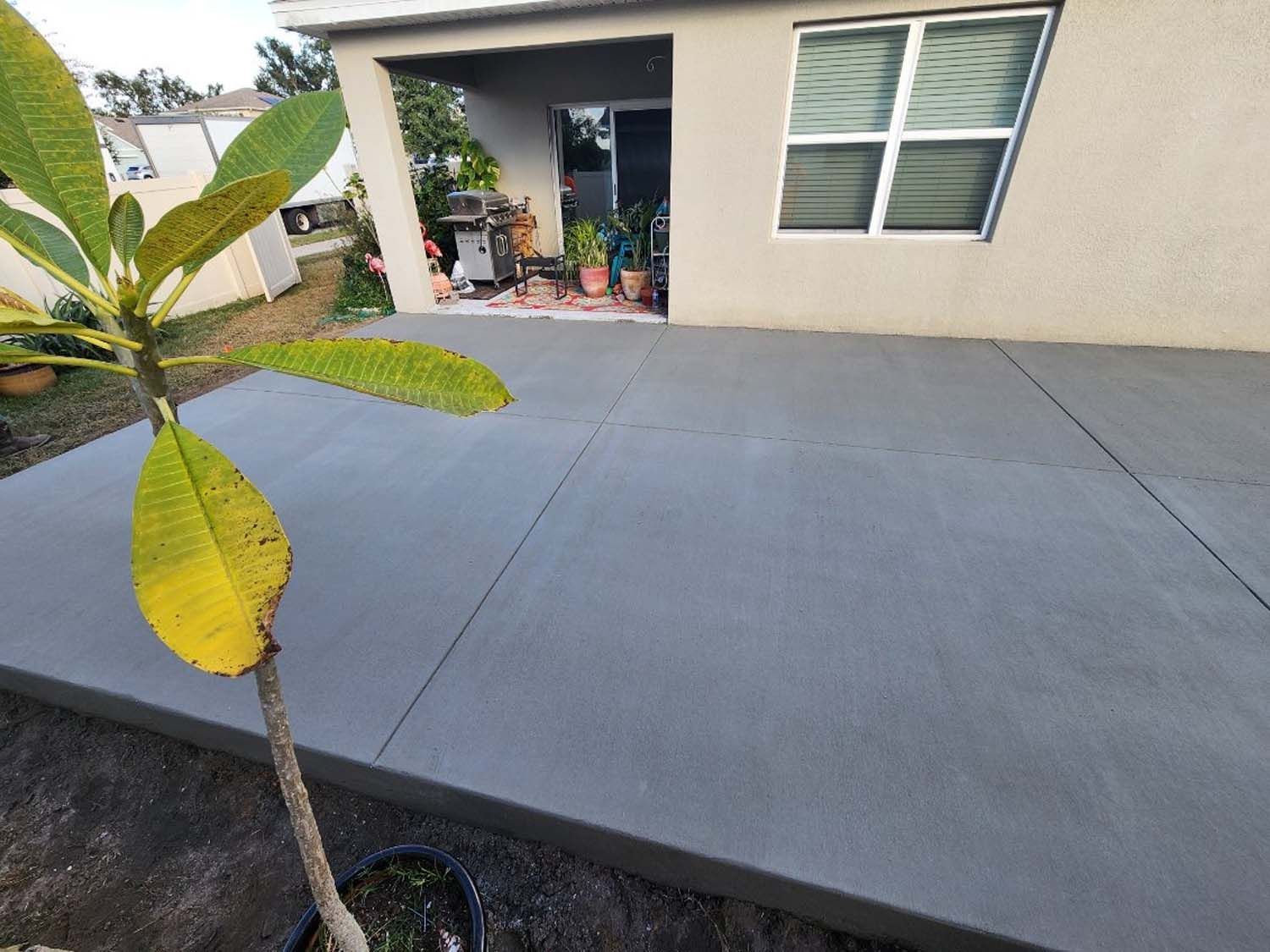 Newly poured gray concrete patio next to a house with a window and sliding door, plants in the background.