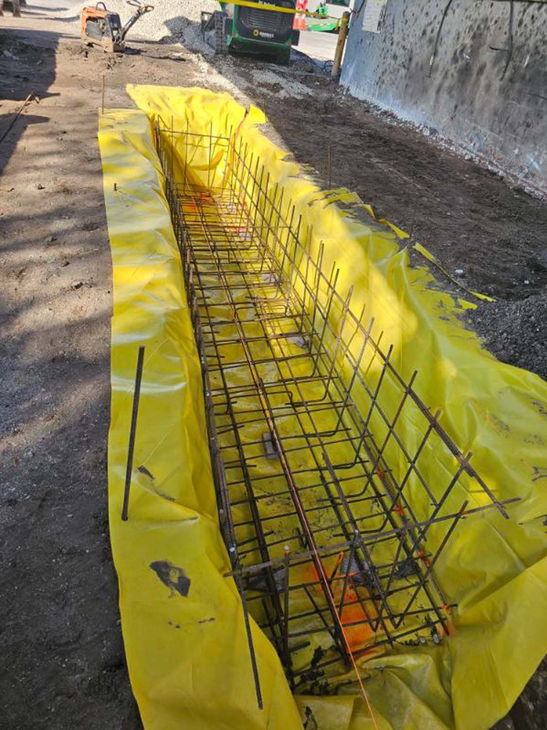 Reinforced steel grid in yellow plastic sheeting, prepared for concrete placement in a construction site trench.