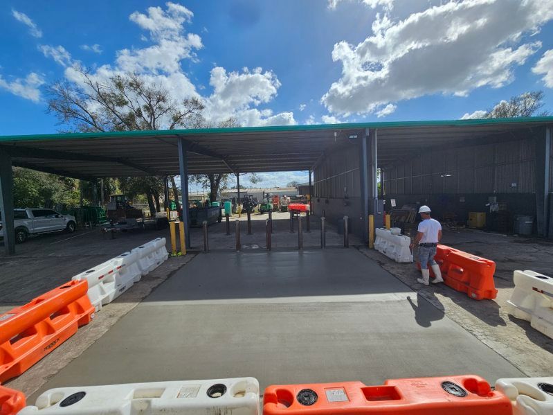 Entrance to a recycling facility with a person, concrete pad, orange/white barriers, and heavy machinery under a canopy. Entrance to a recycling facility with a person, concrete pad, orange/white barriers, and heavy machinery under a canopy.