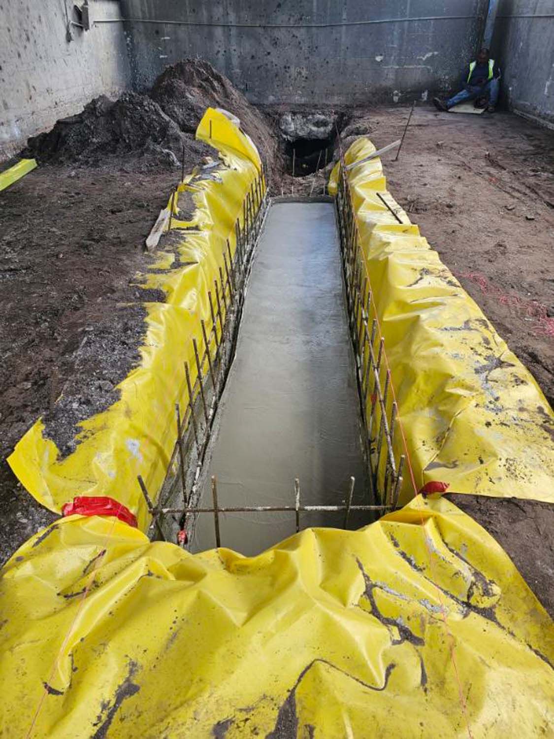 Construction site: concrete trench lined with yellow plastic, rebar visible.