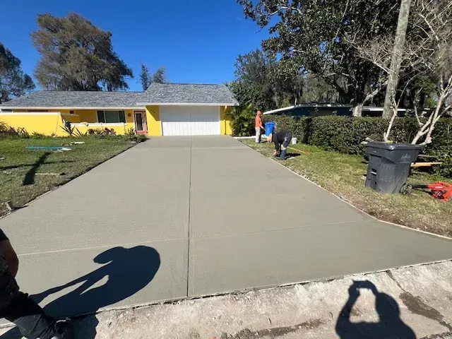 Newly poured concrete driveway in front of a yellow house with a white garage door. Workers are present.