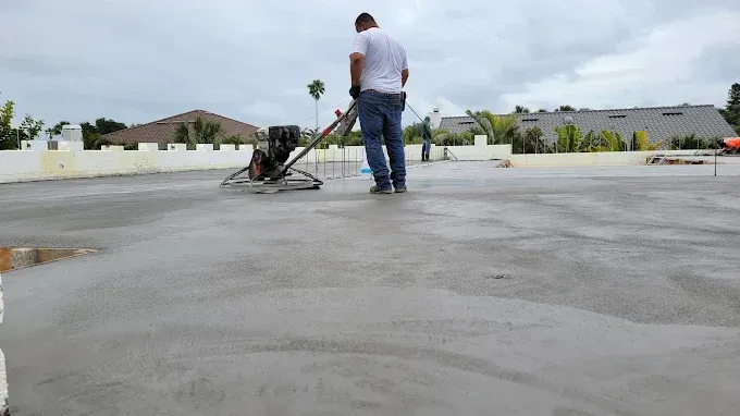 Person wearing orange gloves laying bricks with a trowel, outdoors. Man using a concrete power trowel on a flat, wet concrete surface outdoors.