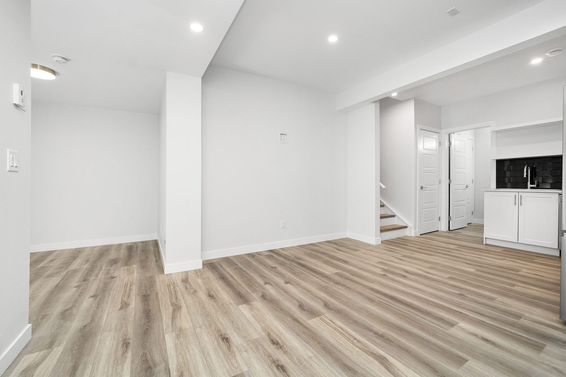 Bright empty modern living room with light wood floors, white walls, and a small kitchen area.
