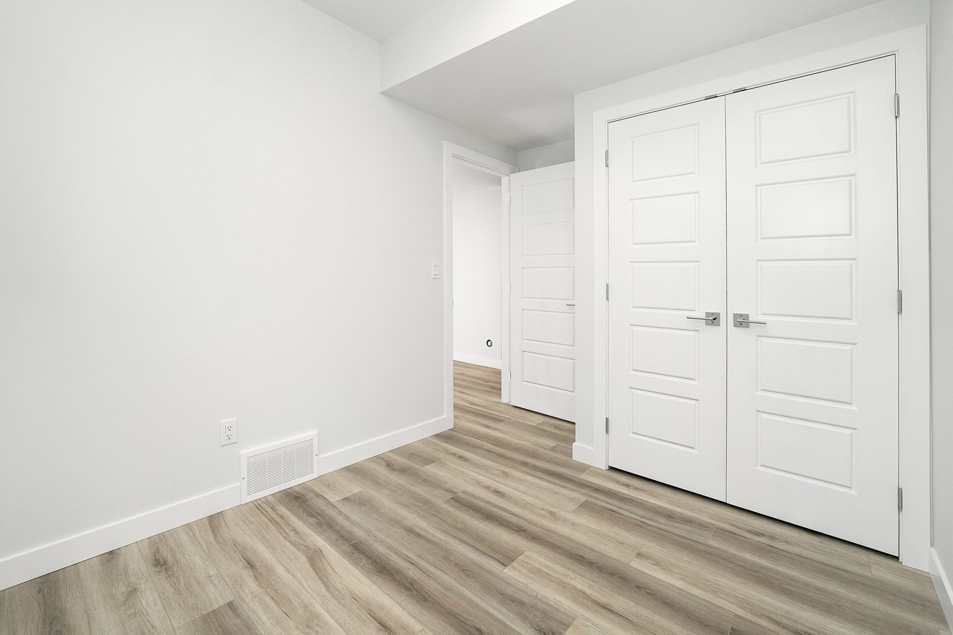 Empty white room with wood flooring and double closet doors near a hallway entrance