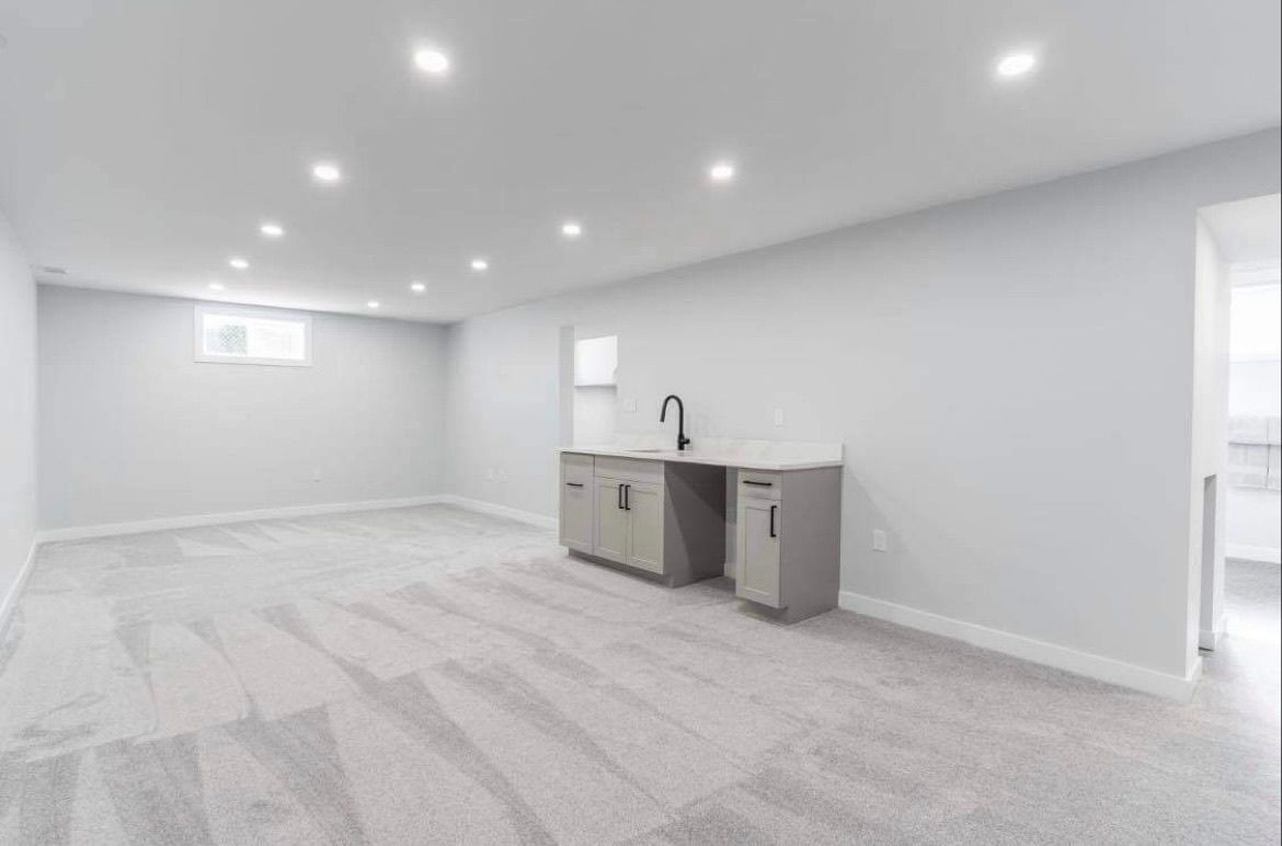 Bright empty basement room with white walls, carpet, recessed lights, and a small wet bar.