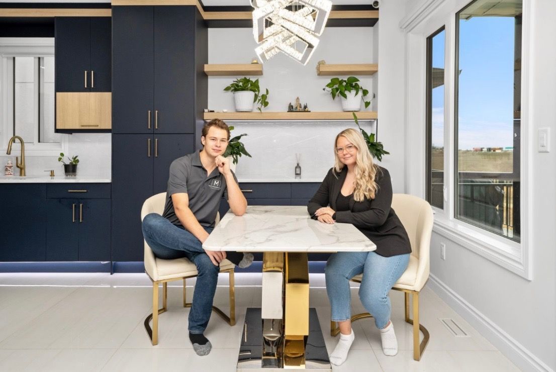 Two people seated at a kitchen table in a bright modern home, smiling with shelves and large windows behind them