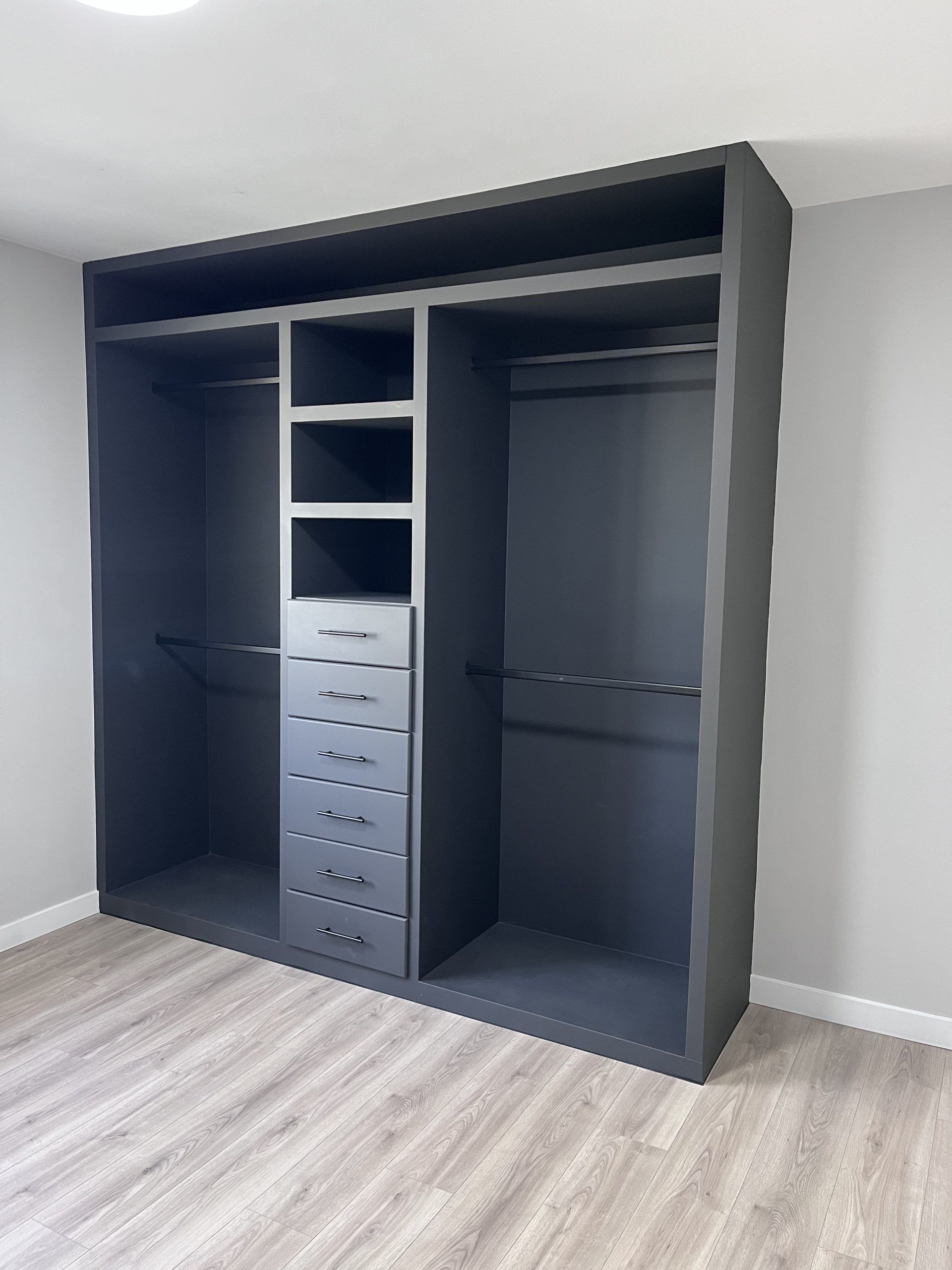 Open dark wardrobe with shelves, hanging space, and drawers in a light gray room.