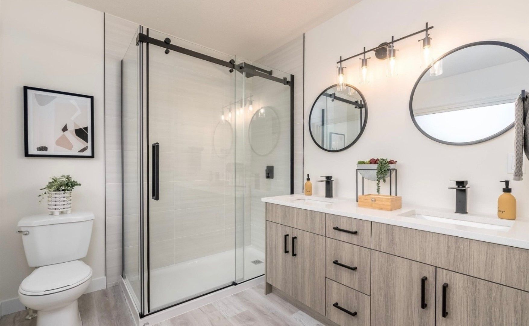 Bright modern bathroom with glass shower, toilet, double vanity, round mirrors, and light wood cabinets.