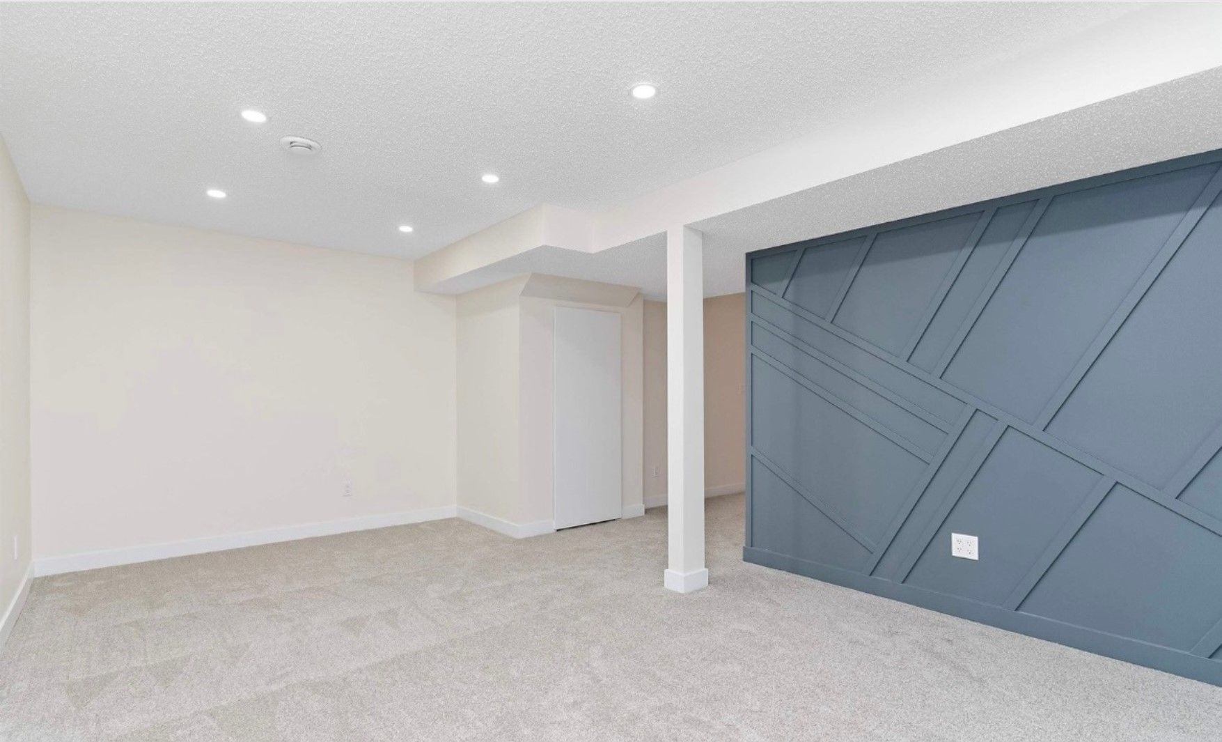 Empty modern basement room with gray carpet, white walls, recessed lights, and a blue accent wall