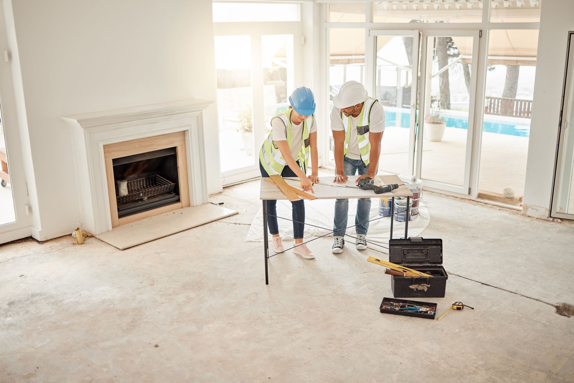 Two workers in hard hats measuring in an unfinished living room with fireplace and sliding glass doors.