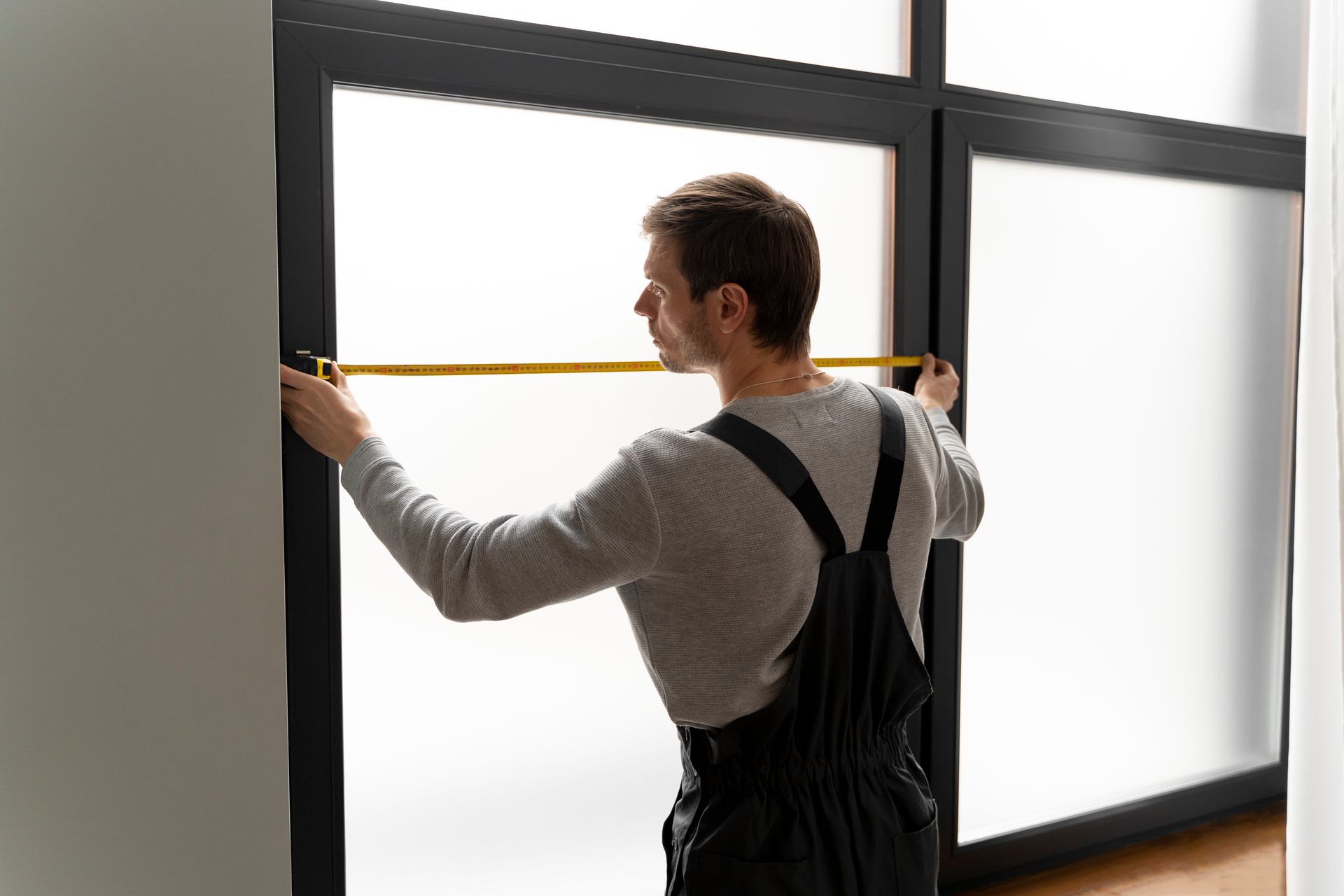 Person measuring a large window frame with a yellow tape measure in a bright room