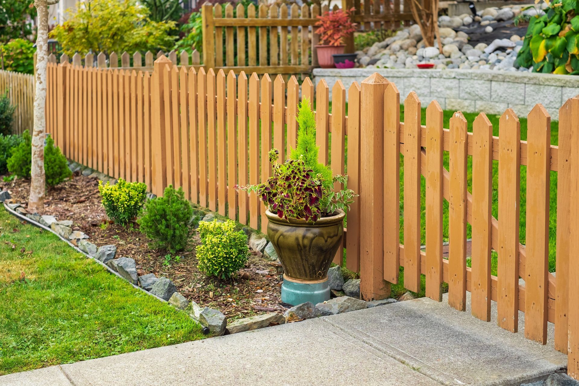 Wooden picket fence along a landscaped garden beside a concrete walkway.