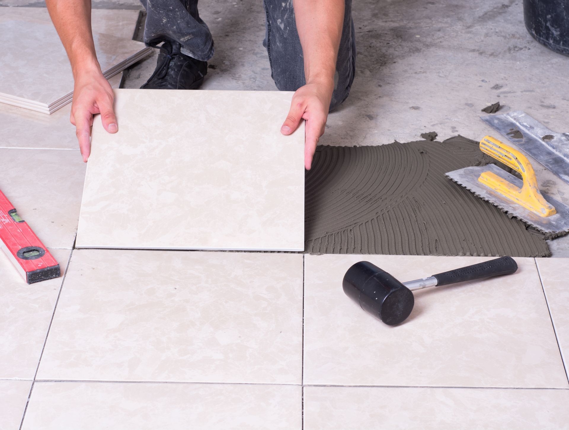 Installing white floor tiles beside a dark adhesive bed with a mallet and trowel nearby
