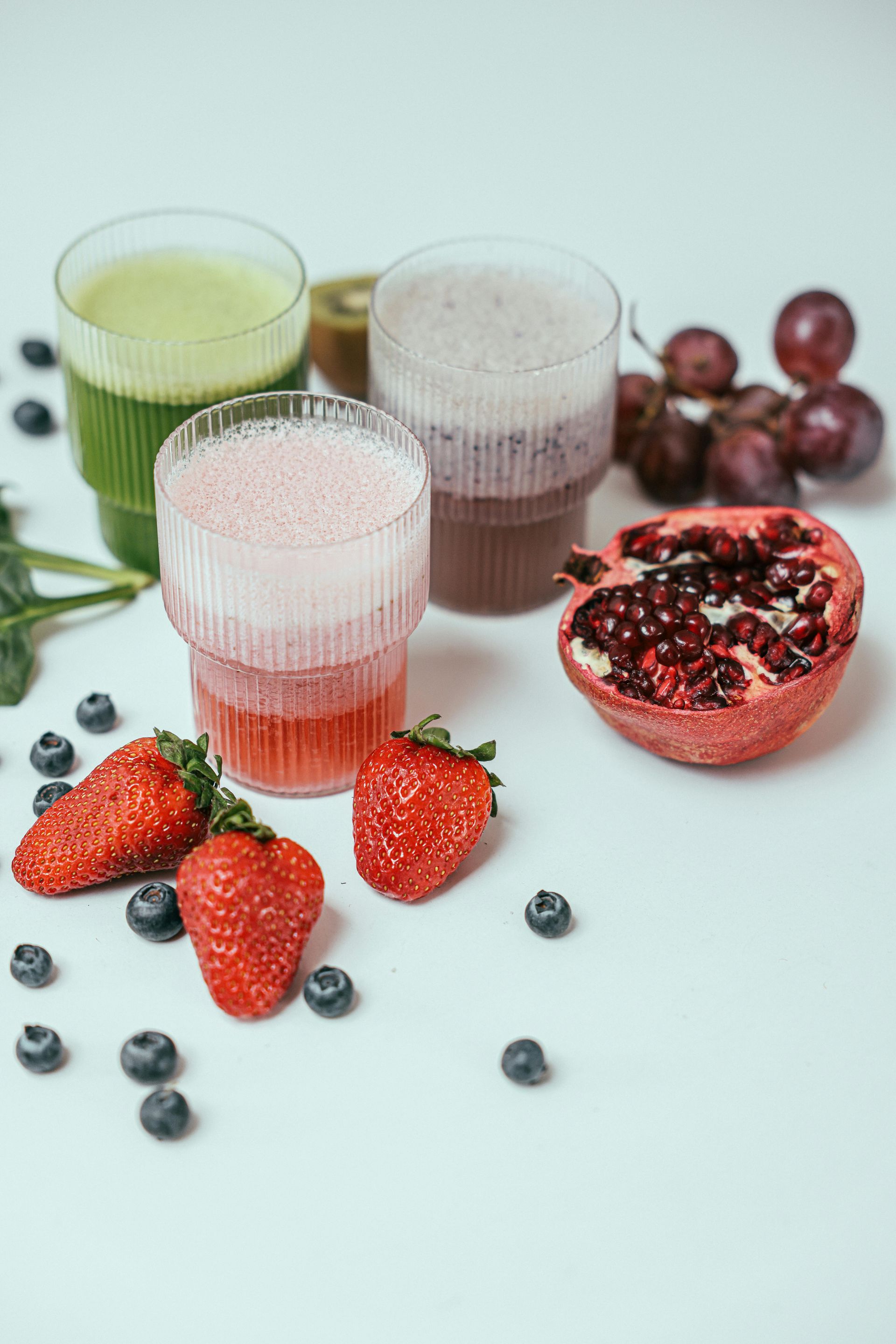 Three glasses of smoothies with strawberries , blueberries , grapes and a pomegranate on a table.