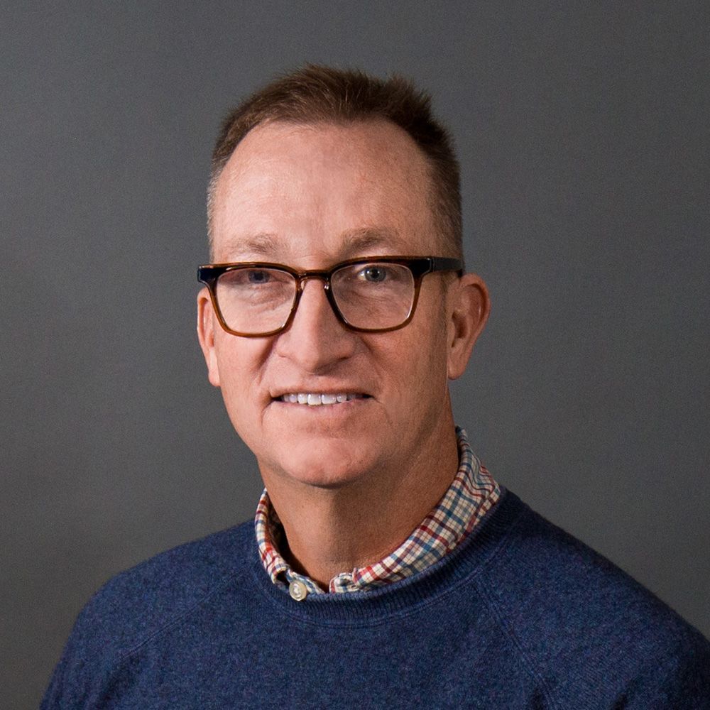 Man wearing glasses, blue sweater, plaid shirt collar against gray backdrop.