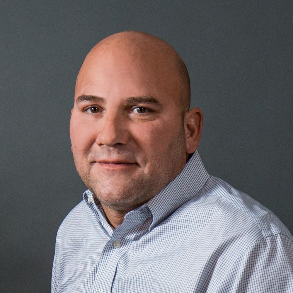 Bald man in a light blue patterned shirt smiles at the camera against a grey backdrop.