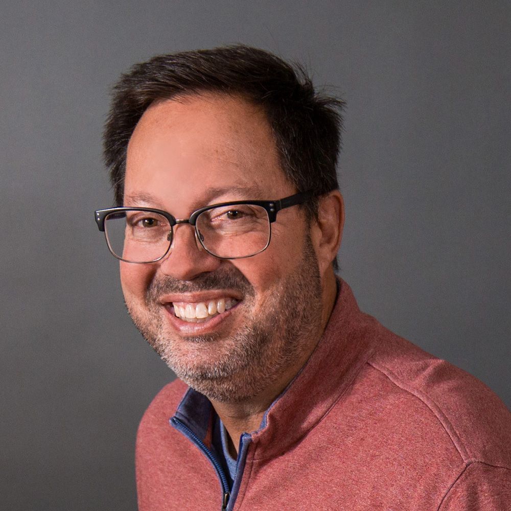 Man with glasses smiles, wearing a red long-sleeve shirt, against a gray background.