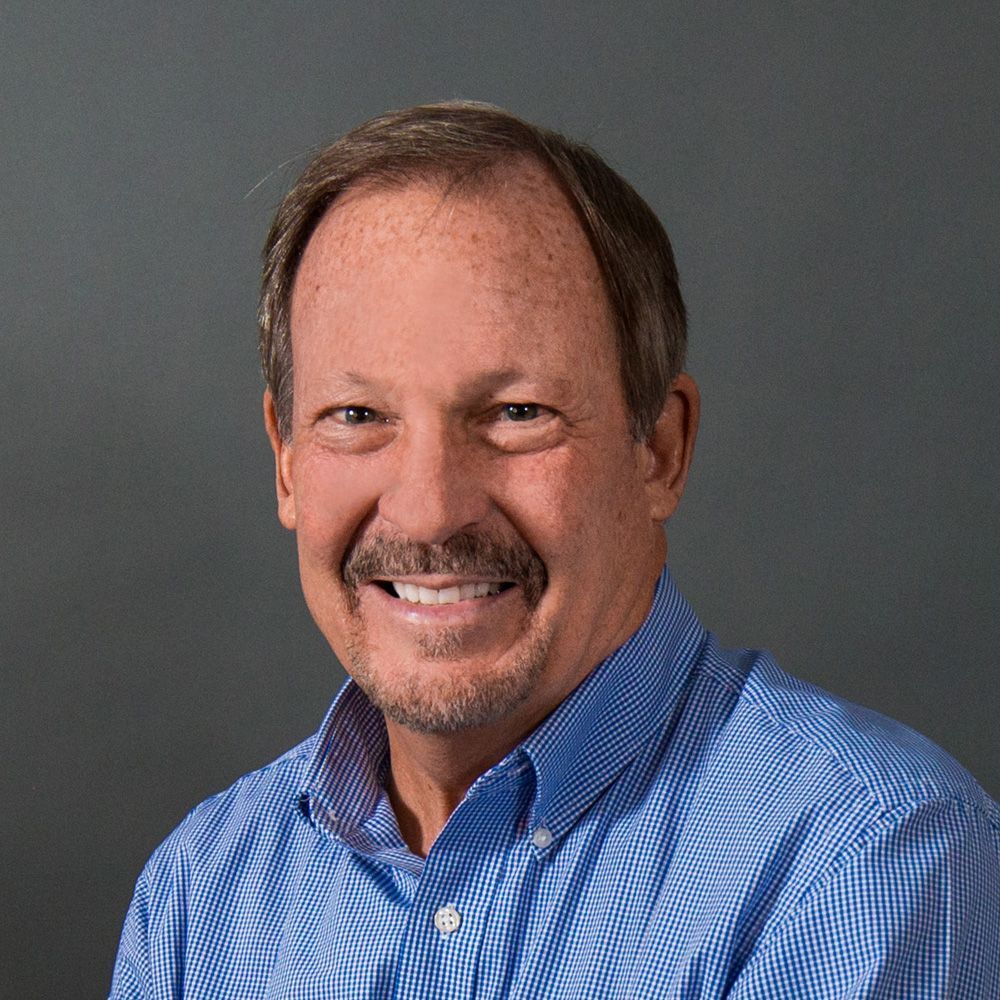 Smiling man with short brown hair, wearing a blue button-down shirt, set against a gray background.