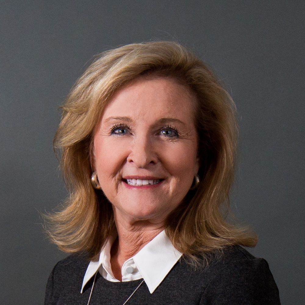 Woman with blonde hair, smiling, wearing a dark blazer over a white collared shirt, against a gray background.