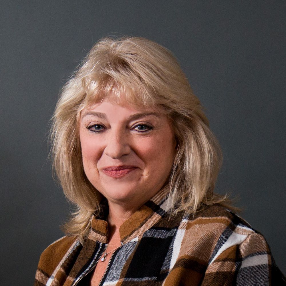 Blonde woman with a slight smile, wearing a brown plaid jacket, against a gray backdrop.