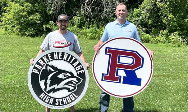 Two men holding round school logos outdoors. One holds a black & white wolf logo, the other a red, white, & blue 