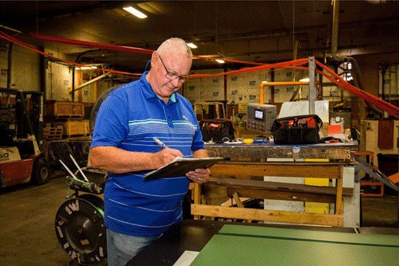 Man in blue shirt, glasses, writing on a clipboard in a warehouse setting, near machinery and equipment.