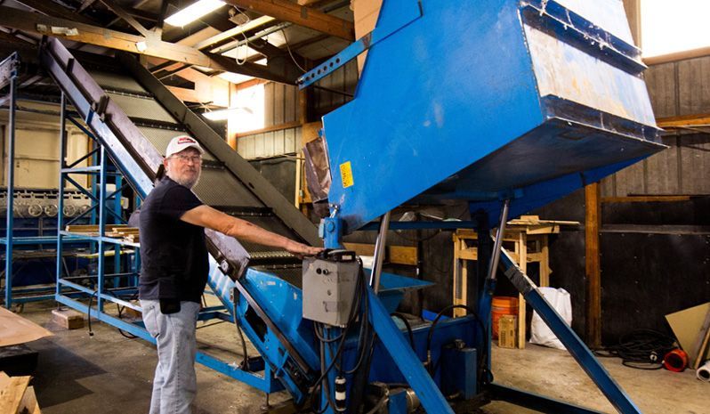 Man standing by a large blue industrial machine with a conveyor belt in a workshop.
