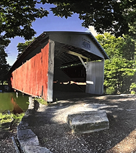 Red covered bridge over water with gray stone foundation, framed by green trees.