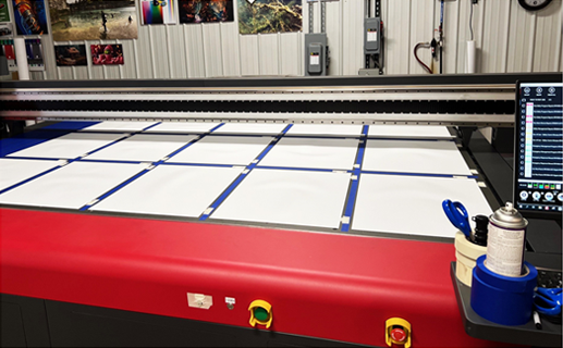 Large format printer with white paper and blue tape grid. Red machine, monitor, and spray can.