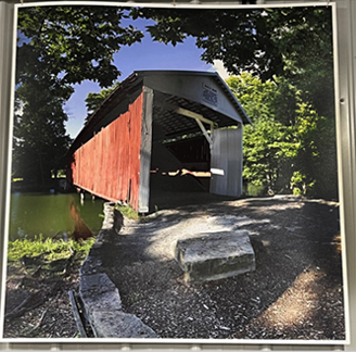 Red and gray covered bridge over water, surrounded by trees under a blue sky.