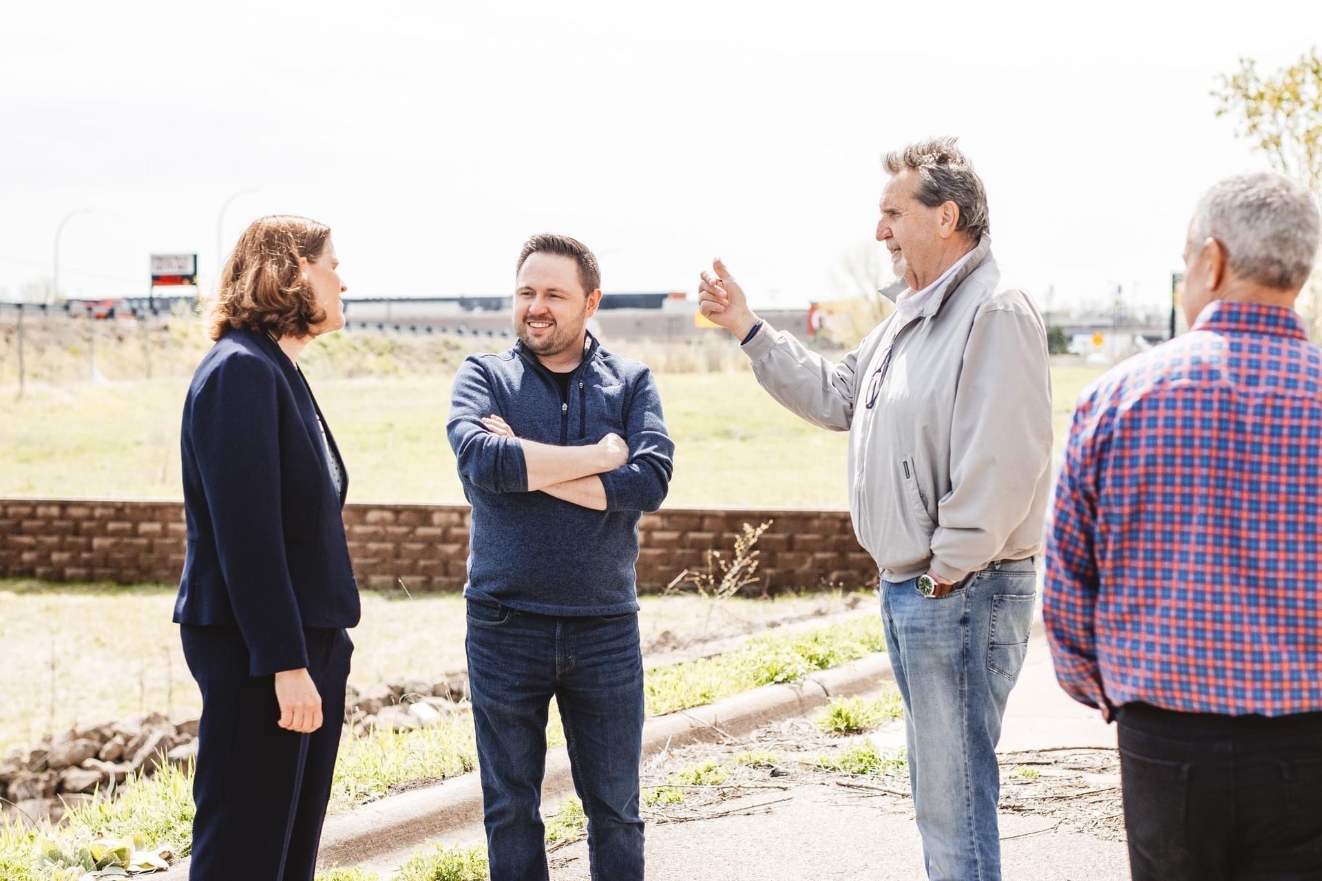 Four people in casual attire converse outdoors; one gestures, others listen.