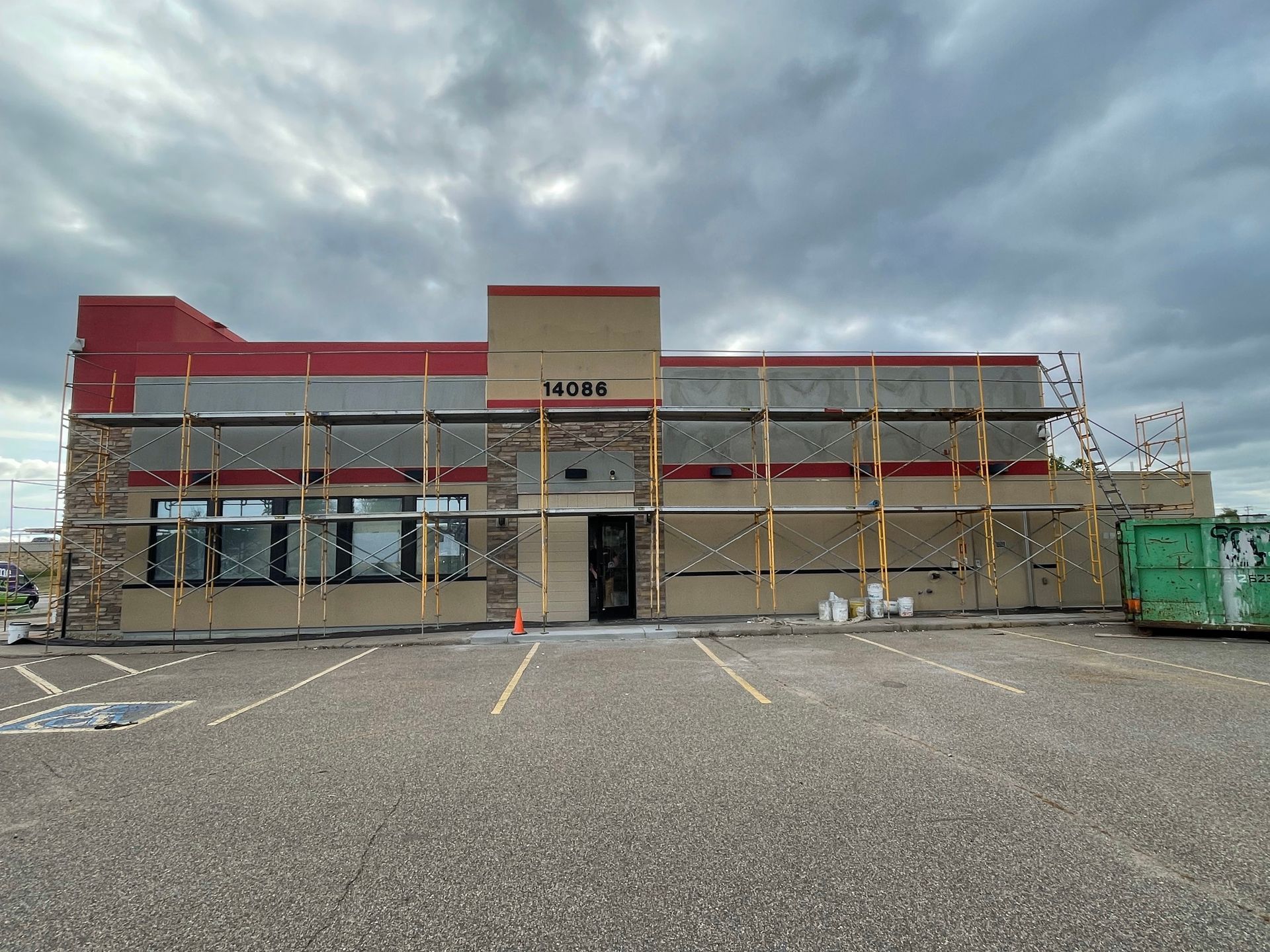 Building exterior under construction with scaffolding, beige walls, red trim, and overcast sky.