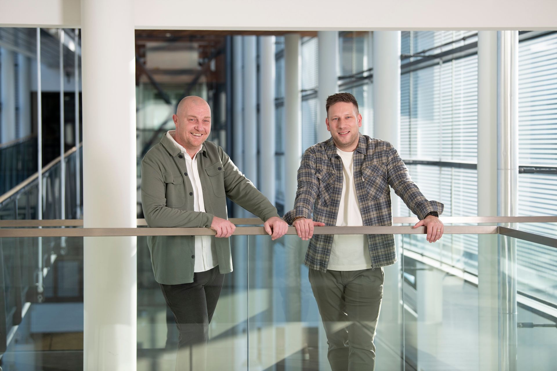 A smiling professional sits at a desk with clasped hands, viewed through a glass partition in a blurred office setting.