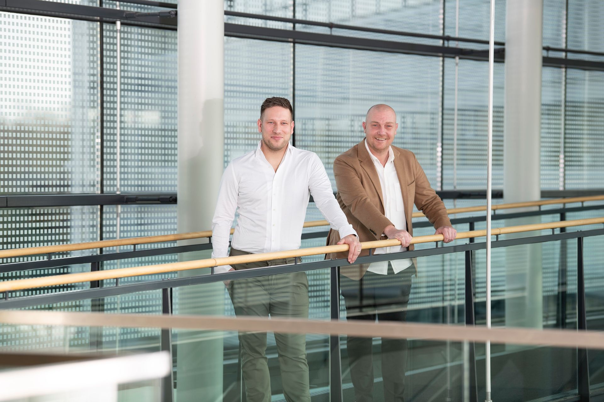 A person in a beige sweater sits at a wooden table, looking at a smartphone while a laptop is open in front of them.
