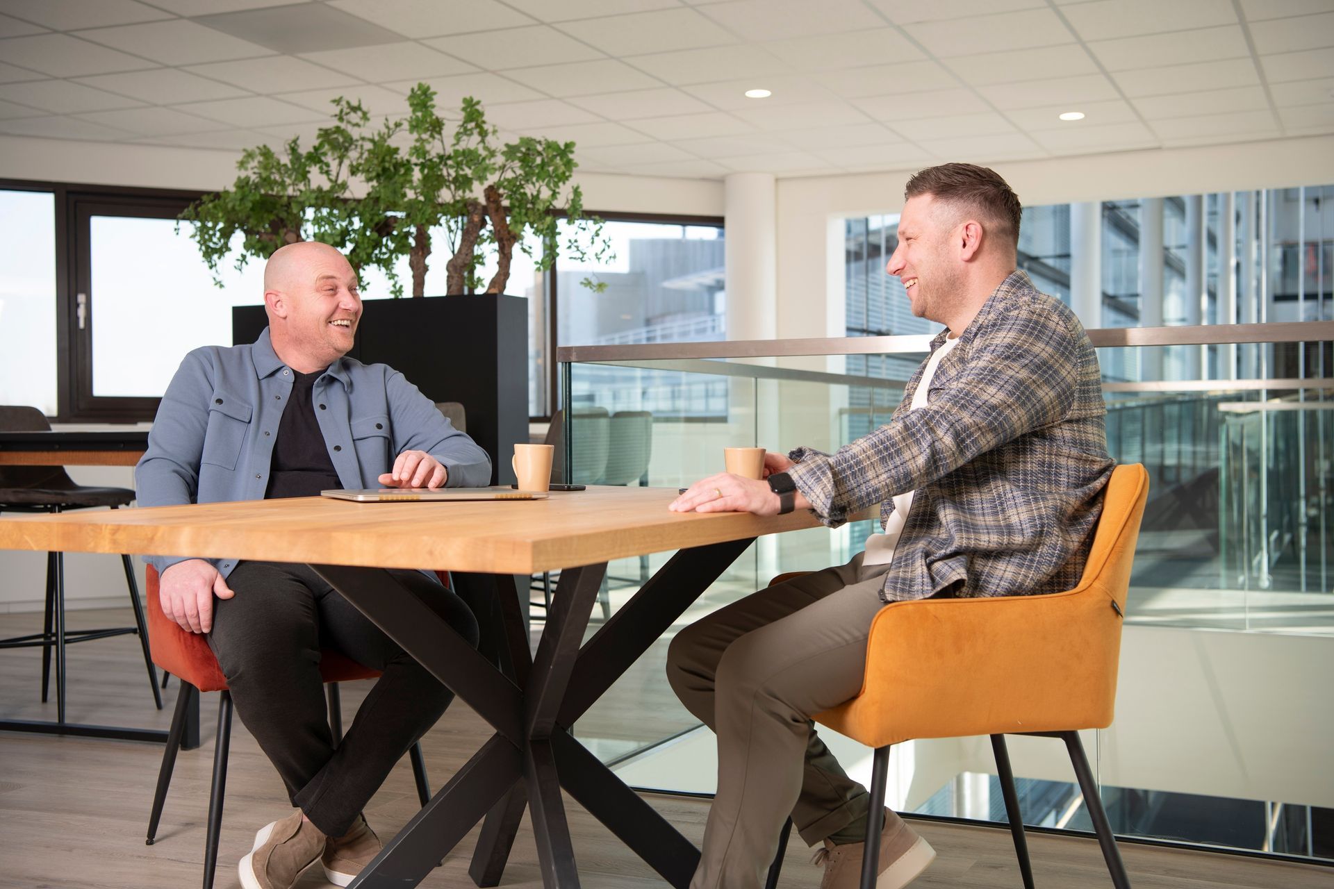 A person in a beige sweater sits at a wooden table, looking at a smartphone while a laptop is open in front of them.