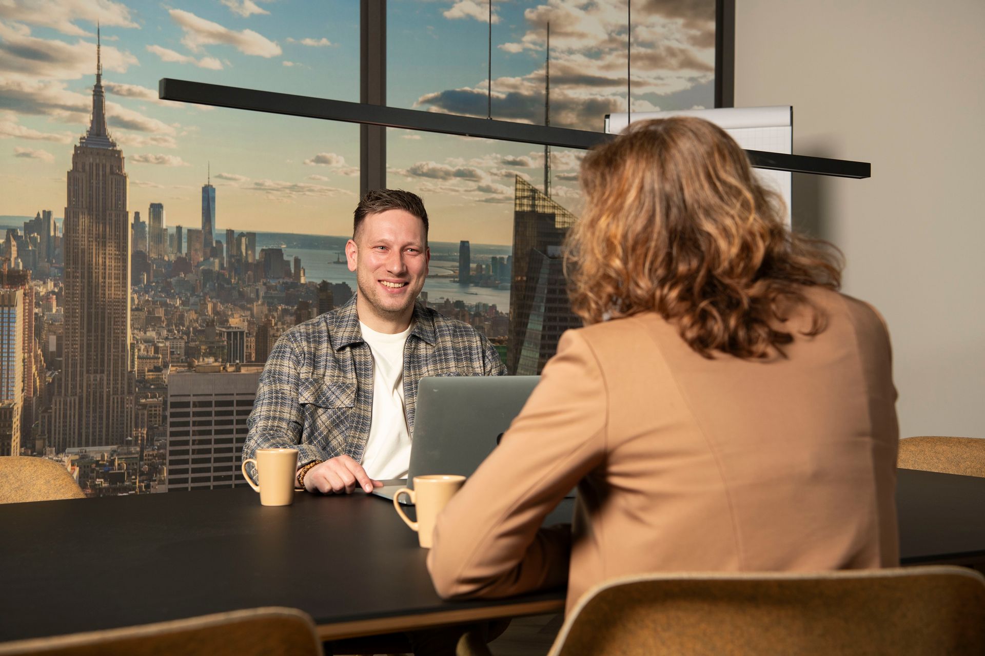A person with reddish hair sits at a wooden table by a window, reading a document in a dimly lit, cozy indoor space.